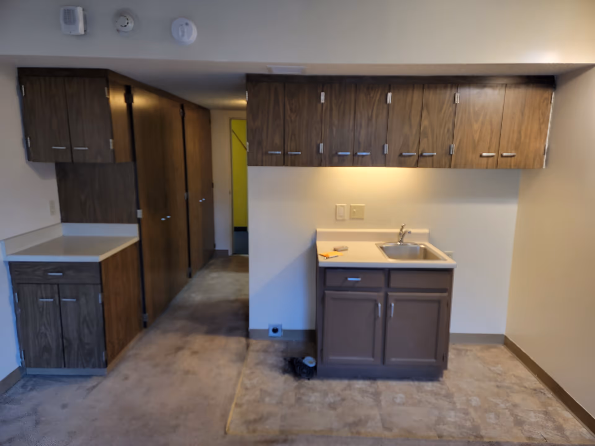Interior view of a small kitchen area in a senior housing residence featuring wooden cabinets above and below a countertop with a sink. The floor transitions from carpet to tile near the kitchen space. There is a hallway with additional wooden cabinets and doors leading further into the residence.