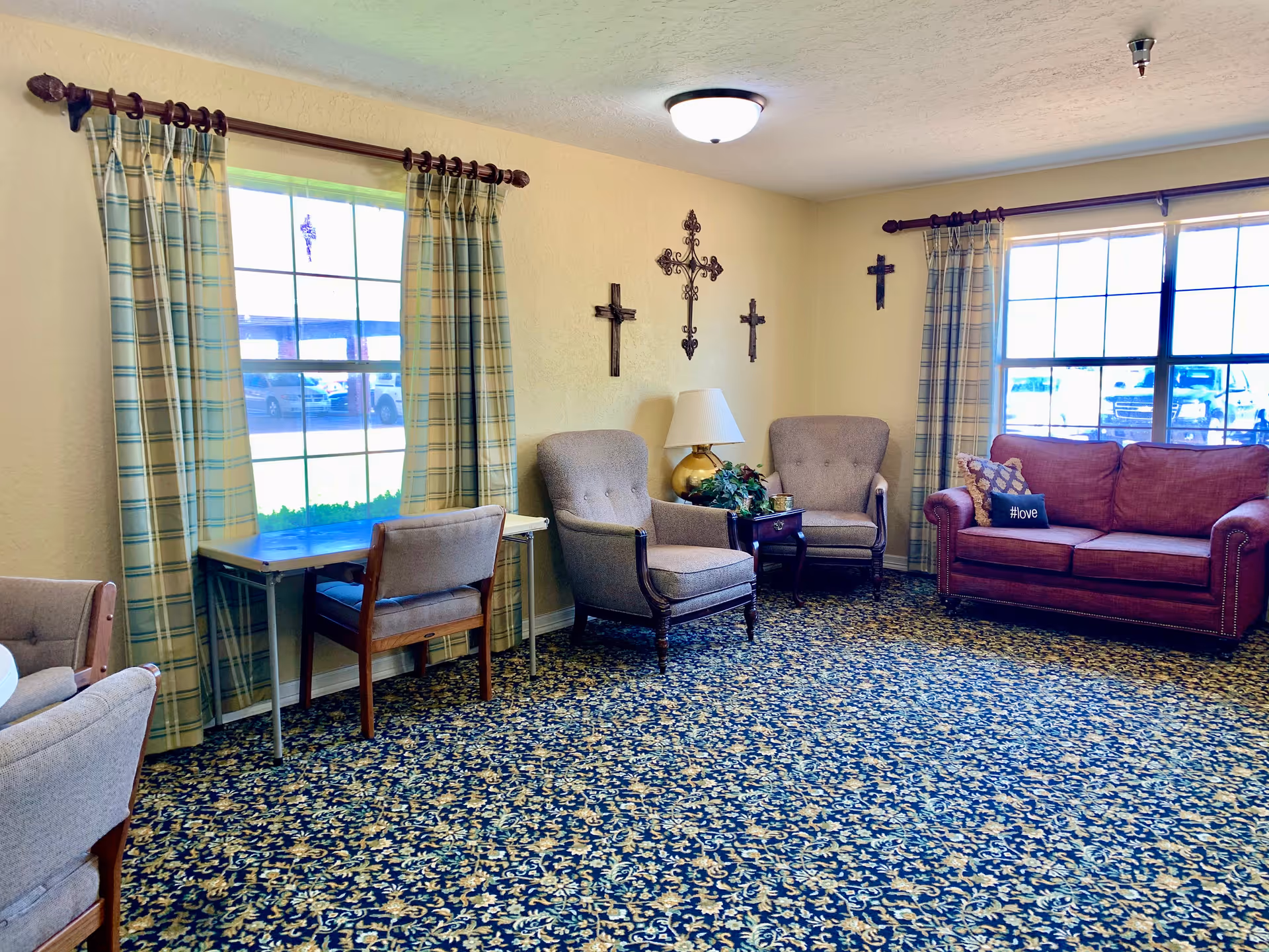 A cozy living room area in a senior living facility with patterned carpet, two large windows with plaid curtains, two beige armchairs, a red sofa with a #love pillow, a small table with a lamp and plant, and decorative crosses on the wall.
