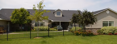 Exterior view of a single-story residential building with beige siding, a dark roof, and several windows. The building is surrounded by a well-maintained lawn, trees, shrubs, and a black metal fence in the foreground.