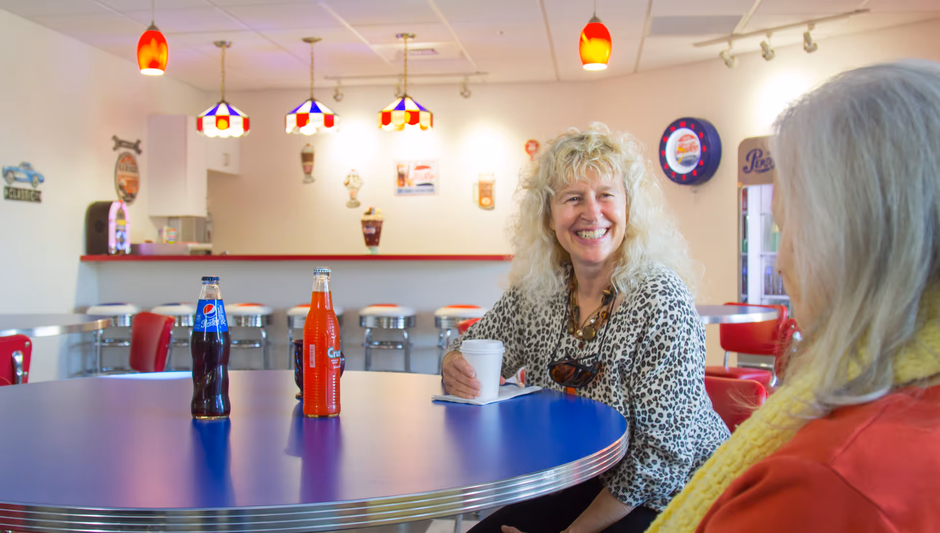 Two women sitting at a round blue table in a retro-style diner or cafe area. One woman with curly blonde hair and a leopard print top is smiling and holding a white cup. The other woman, with long gray hair and a yellow scarf, is facing her. On the table are two glass soda bottles, one Pepsi and one Crush. The background features red and white bar stools, vintage-style hanging lamps, and retro wall decorations.