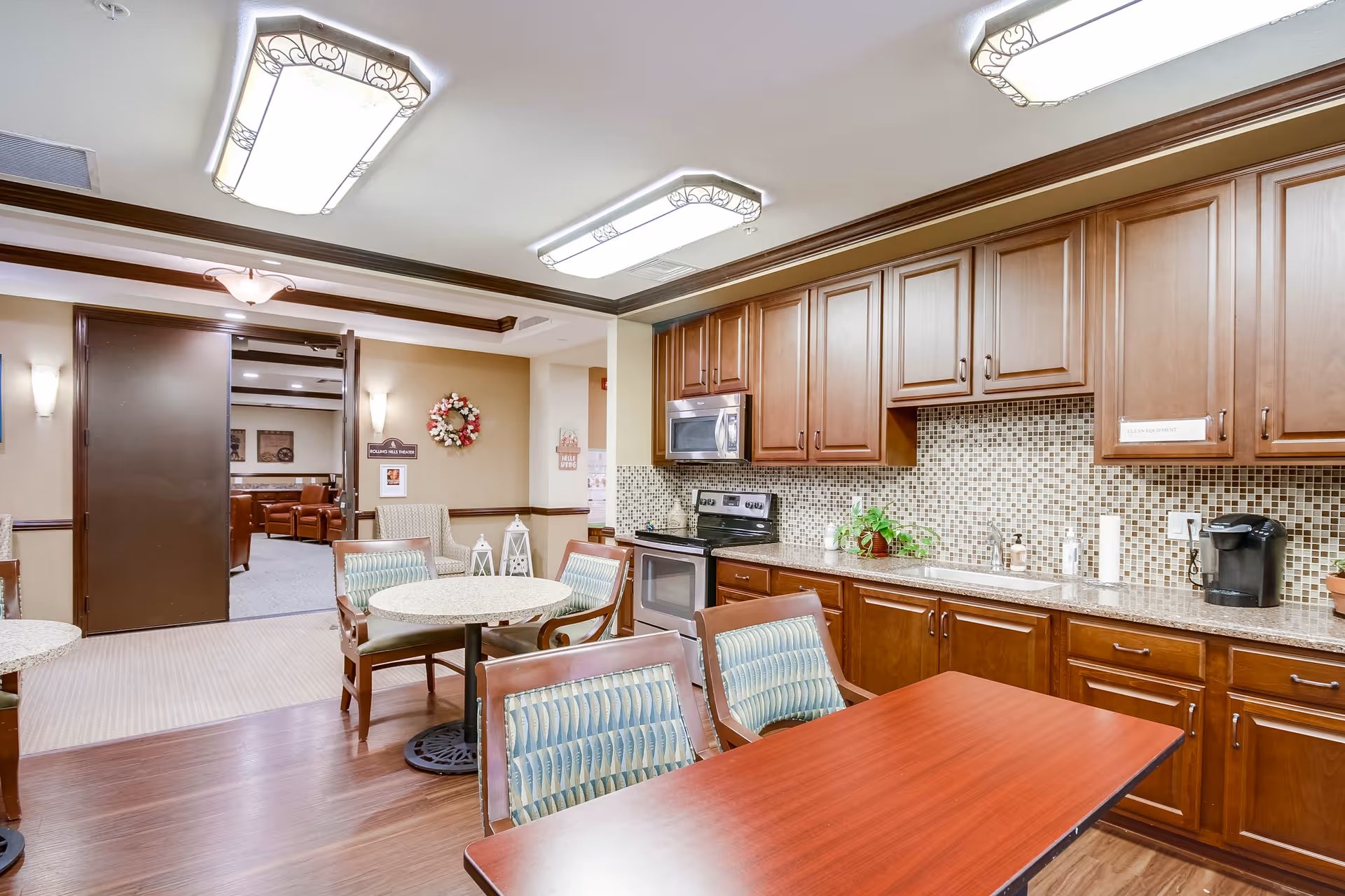 Interior view of a senior living facility kitchen and dining area with wooden cabinets, a mosaic tile backsplash, stainless steel appliances, and several tables with chairs. The room is well-lit with decorative ceiling lights, and an open doorway leads to a lounge area with brown leather seating.