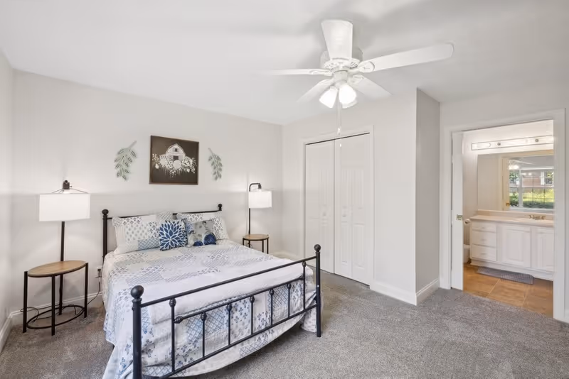 A bedroom with a black metal bed frame, patterned bedding in shades of blue and white, two bedside tables with lamps, a ceiling fan with lights, a closet with double doors, and an open doorway leading to a bathroom with a large mirror and white cabinetry.