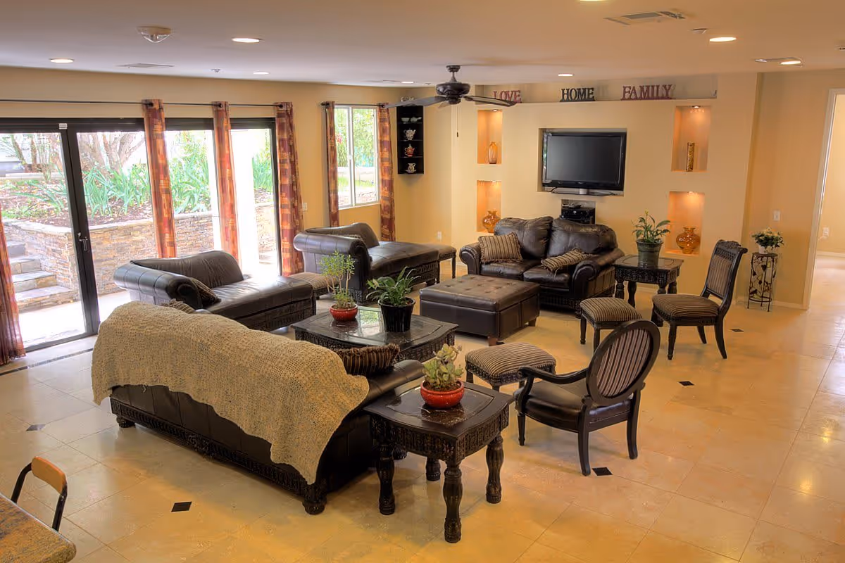 A spacious living room with multiple leather sofas and chairs arranged around a central coffee table. The room has large windows and glass doors with curtains, allowing natural light to enter. There is a wall-mounted TV above a built-in shelf with decorative items and the words 'LOVE', 'HOME', and 'FAMILY' displayed above it. The floor is tiled, and there are several potted plants on tables throughout the room.