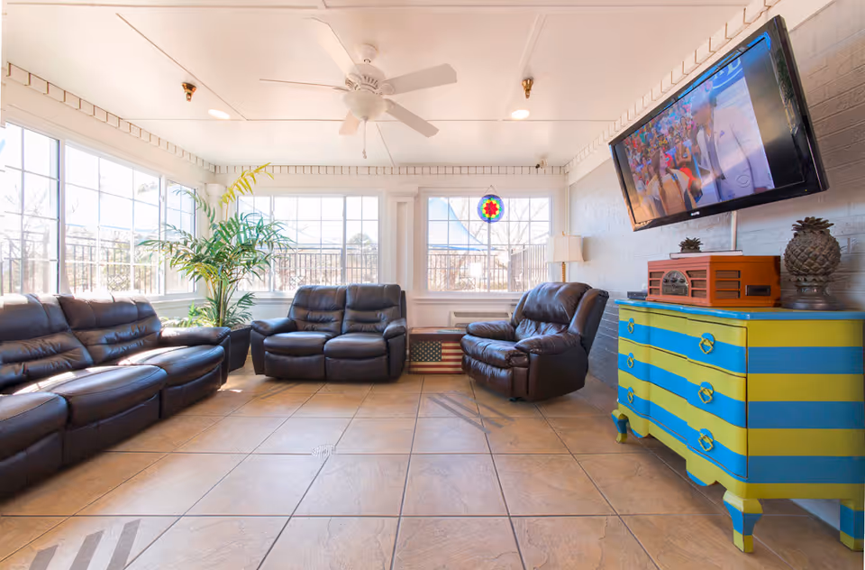 A bright and spacious living room with large windows letting in natural light. The room features three black leather recliner sofas arranged around the space. There is a colorful blue and yellow striped dresser with decorative items on top, including a vintage-style radio and two pineapple-shaped ornaments. A flat-screen TV is mounted on the wall above the dresser. The floor is tiled, and a ceiling fan is installed overhead. A green potted plant is placed near the windows.