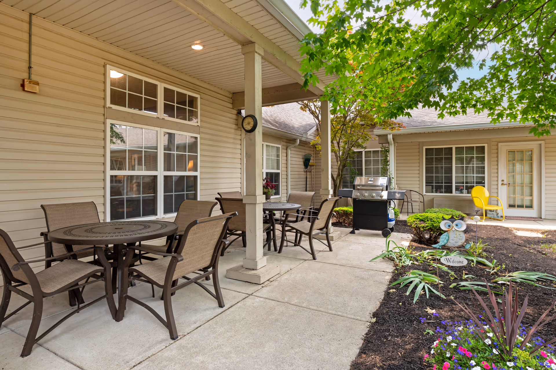 Outdoor patio area at Brookdale Richmond with round tables and chairs under a covered porch. There is a barbecue grill, a yellow chair near a door, and a garden with flowers and a decorative owl statue holding a welcome sign. Trees provide shade over the area.