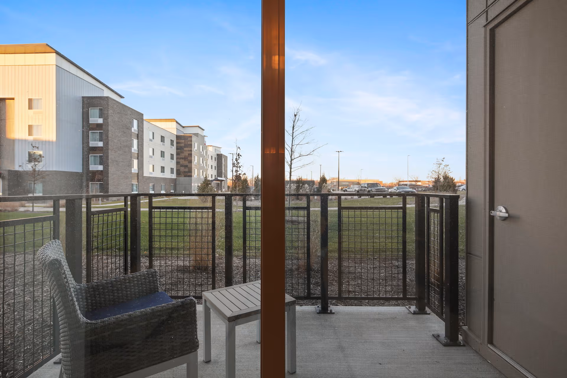 View through a glass door onto a small outdoor patio with a wicker chair and a small table. The patio is enclosed by a black metal railing. Beyond the patio is a grassy area with a few small trees and a multi-story building in the background under a clear blue sky.