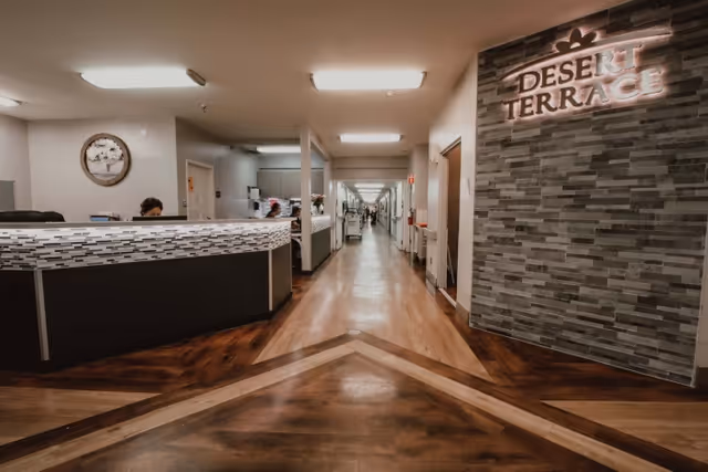 Interior view of a healthcare facility hallway with a reception desk on the left and a decorative wall on the right displaying the sign 'Desert Terrace'. The hallway extends into the distance with doors and lighting along the walls.