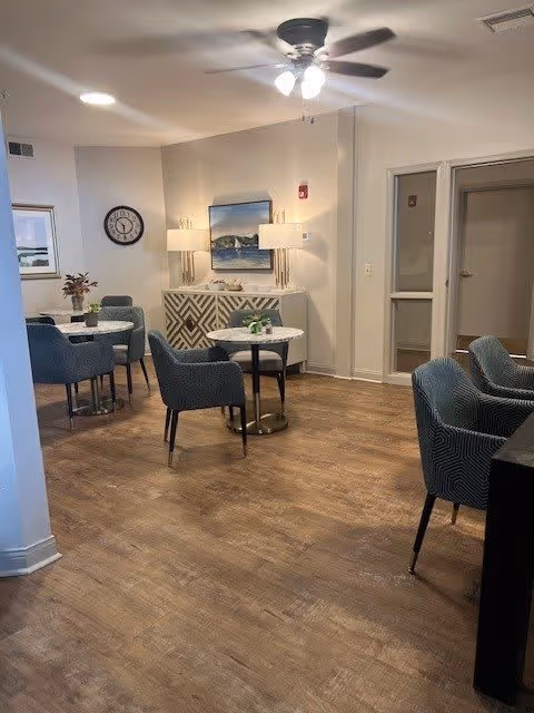 A cozy common area in a senior living facility with wooden flooring, several round tables with blue upholstered chairs, a ceiling fan with lights, a wall clock, a sideboard with lamps and a painting above it, and a doorway leading to another room.