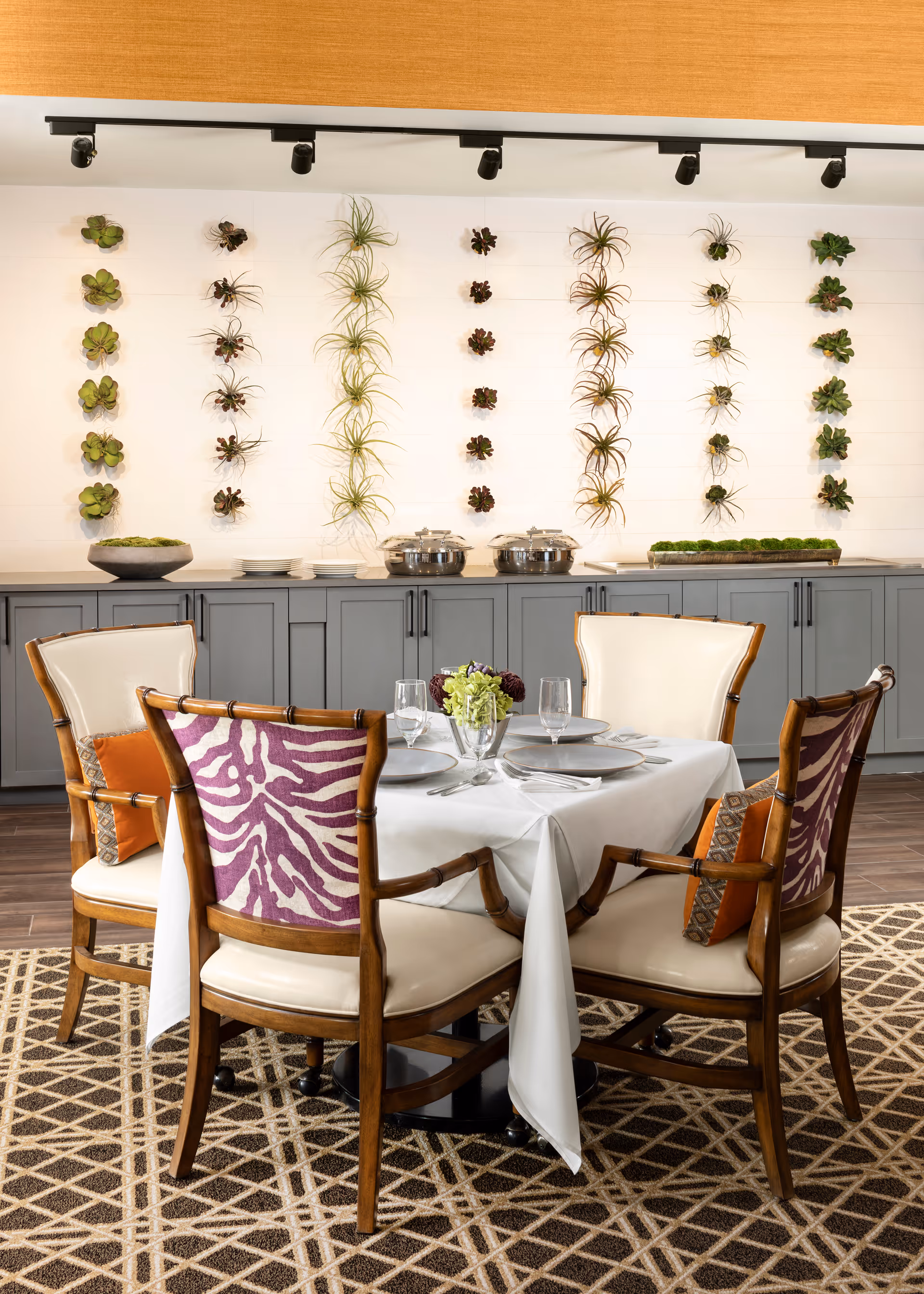 A round dining table set for four with upholstered chairs in a dining room featuring gray buffet cabinets and a decorative wall of small hanging plants.