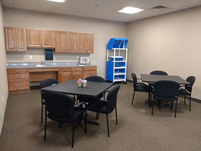 A small dining area with two square black tables, each surrounded by four black chairs. In the background, there is a countertop with a sink, wooden cabinets above and below, and a blue and white utility cart against the wall. The room has beige walls and a carpeted floor.