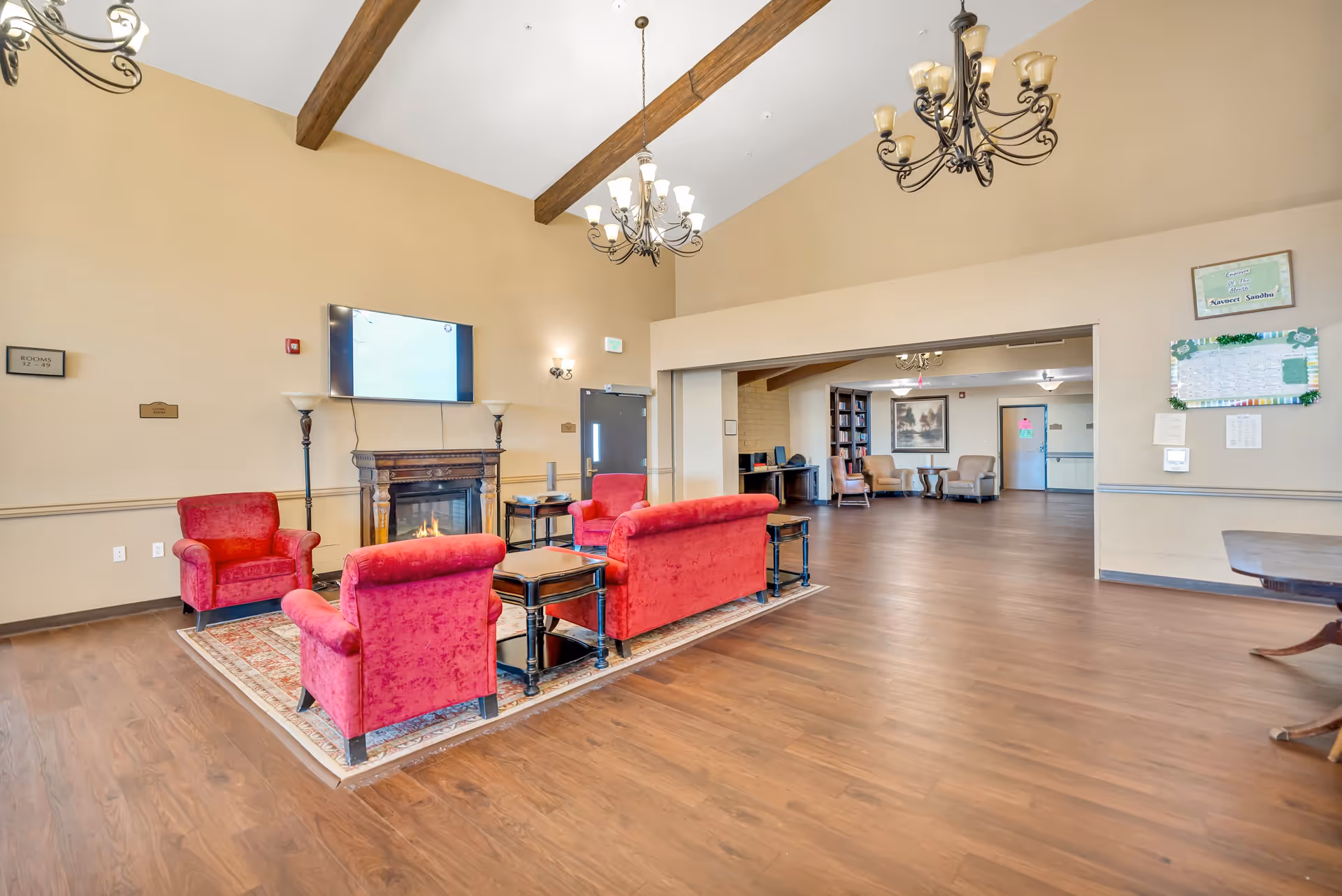 A spacious living room area with wooden floors and high ceilings featuring exposed wooden beams. The room has red upholstered armchairs and sofas arranged around a small wooden table on a patterned rug. A fireplace with a mounted flat-screen TV above it is visible on one wall. Two chandeliers hang from the ceiling, and there is an open doorway leading to another room with additional seating and bookshelves.