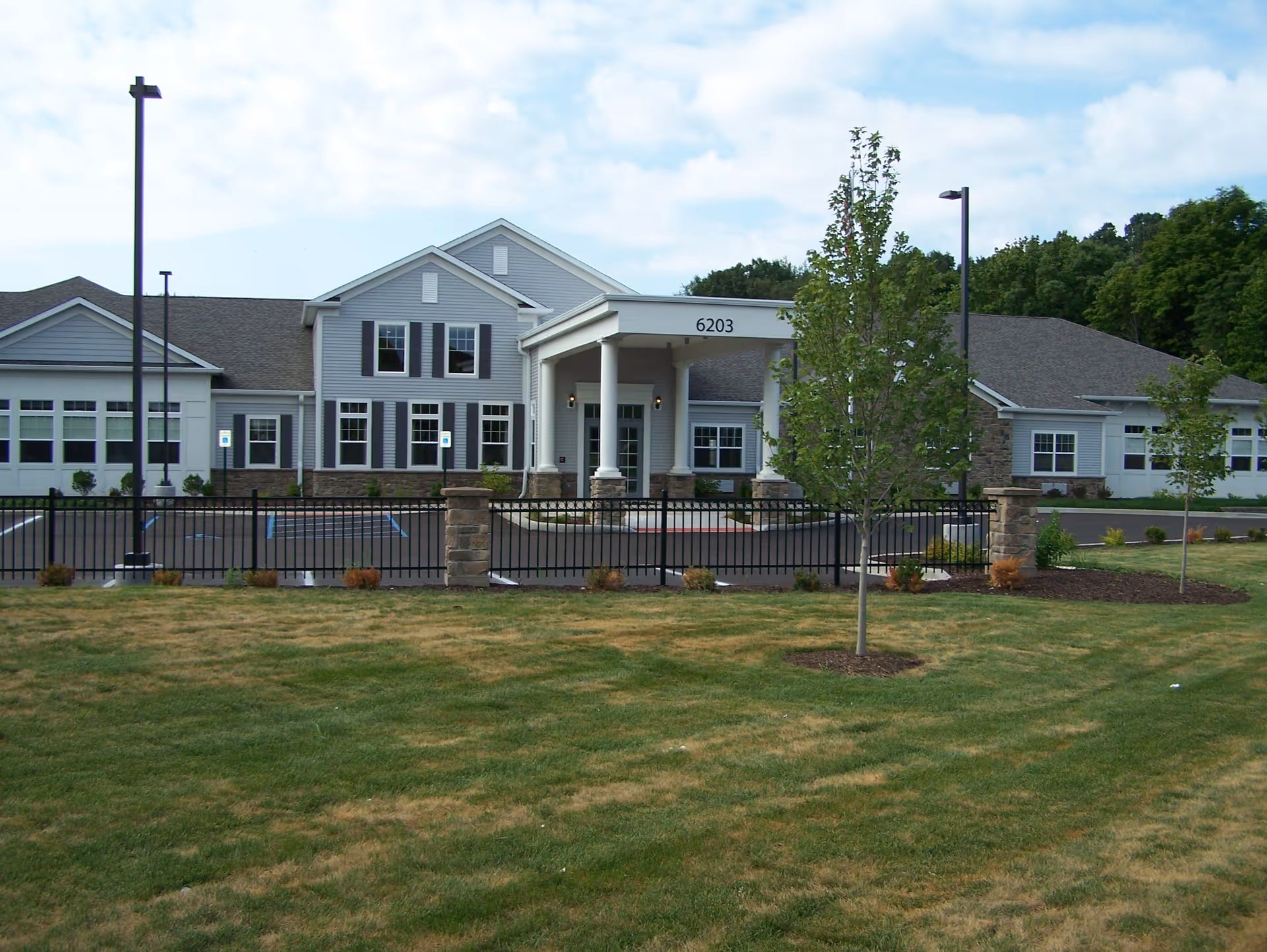 Exterior front view of a single-story building with gray siding and white trim, featuring a covered entrance with columns and the number 6203 above it. There is a black metal fence and a parking lot with handicap parking spaces in front, along with a grassy area and young trees in the foreground.