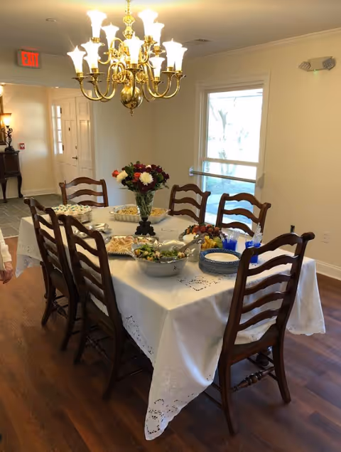 A dining room with a long table covered in a white lace tablecloth, set with bowls of food and a floral centerpiece beneath a brass chandelier.