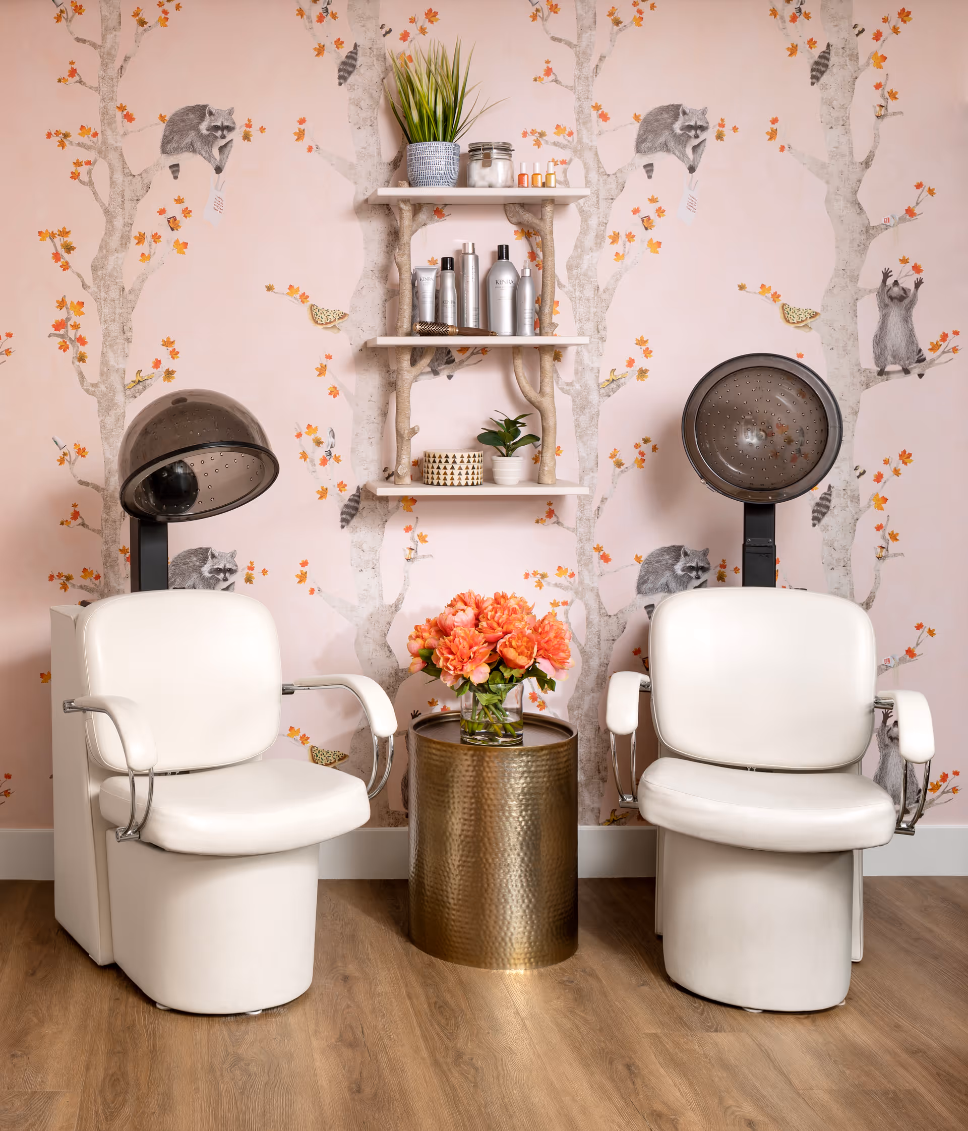 Two white salon dryer chairs flanking a brass side table with flowers against pink tree-and-raccoon wallpaper and shelves holding hair products.