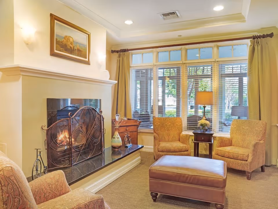 Cozy living room with a lit fireplace protected by a decorative metal screen, two upholstered armchairs, a leather ottoman, a side table with a lamp and flowers, large windows with blinds and curtains, and a framed painting above the fireplace.