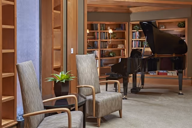 A cozy interior room featuring two upholstered armchairs with wooden arms, a small glass-top side table with a potted plant, a black grand piano, and wooden bookshelves filled with books and decorative plants.