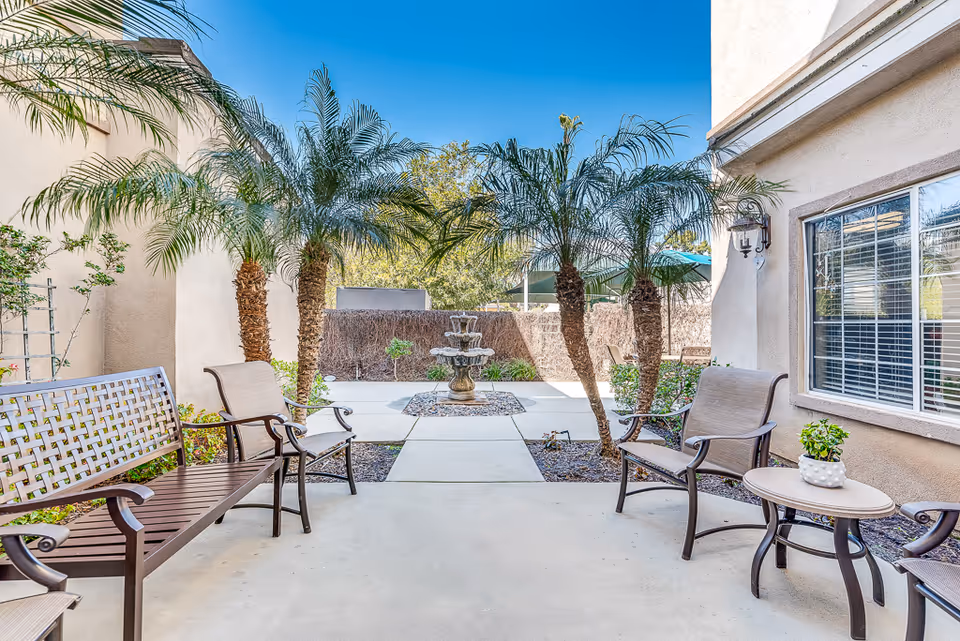 Sunlit courtyard patio with benches and chairs, potted plants and palm trees flanking a central fountain between building walls.