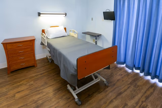 A small patient room with a single hospital bed covered with a gray blanket and a white pillow. Next to the bed is a wooden nightstand with three drawers. A small wall-mounted TV is positioned near the bed, and blue curtains cover the window. The floor is wood-patterned.
