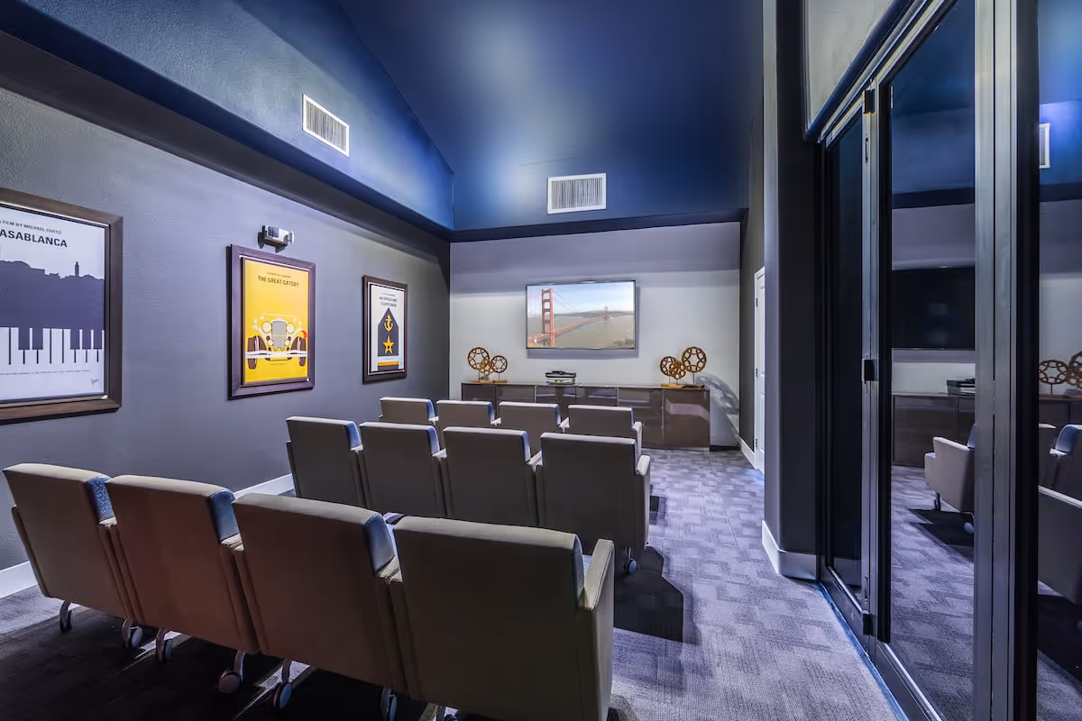 A small theater room with two rows of cushioned chairs facing a wall-mounted flat screen TV displaying an image of the Golden Gate Bridge. The room has dark walls with framed movie posters and decorative film reel sculptures on a cabinet beneath the TV. The floor is carpeted and there is a glass door on the right side.
