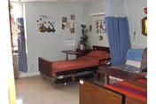 A room in a healthcare facility with two hospital beds, one with a brown blanket and the other with a colorful striped blanket. The room has light-colored walls decorated with framed pictures and wall hangings. There is a blue privacy curtain partially drawn between the beds, a small table with a plant, and a window with curtains letting in natural light.