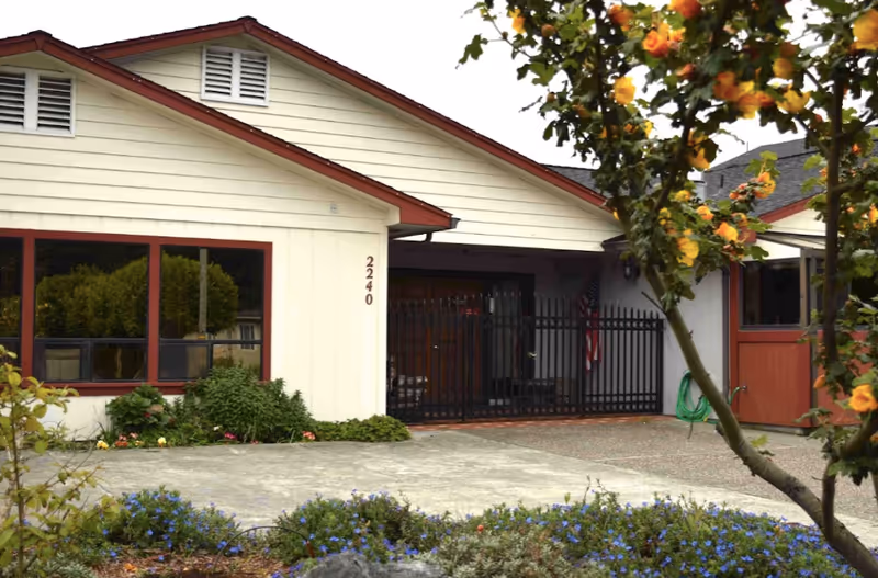 Front exterior of a single-story care home with a gated entrance, driveway, and flowering shrubs.