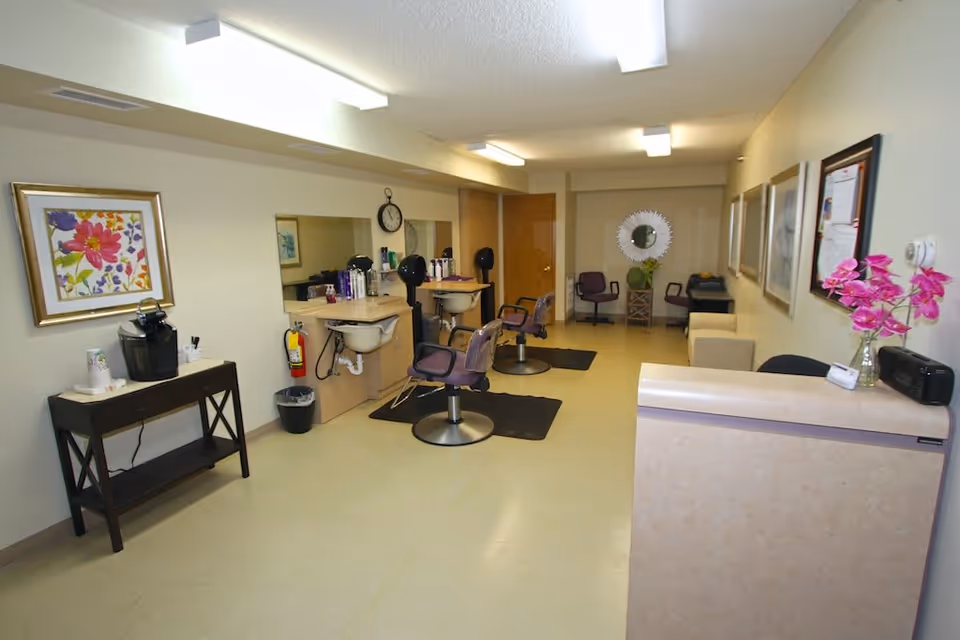 Interior view of a salon area in Heathers Manor featuring two styling chairs in front of mirrors with sinks, a small table with a coffee maker and cups, framed artwork on the walls, a clock, and additional seating in the background. The room is well-lit with fluorescent ceiling lights.