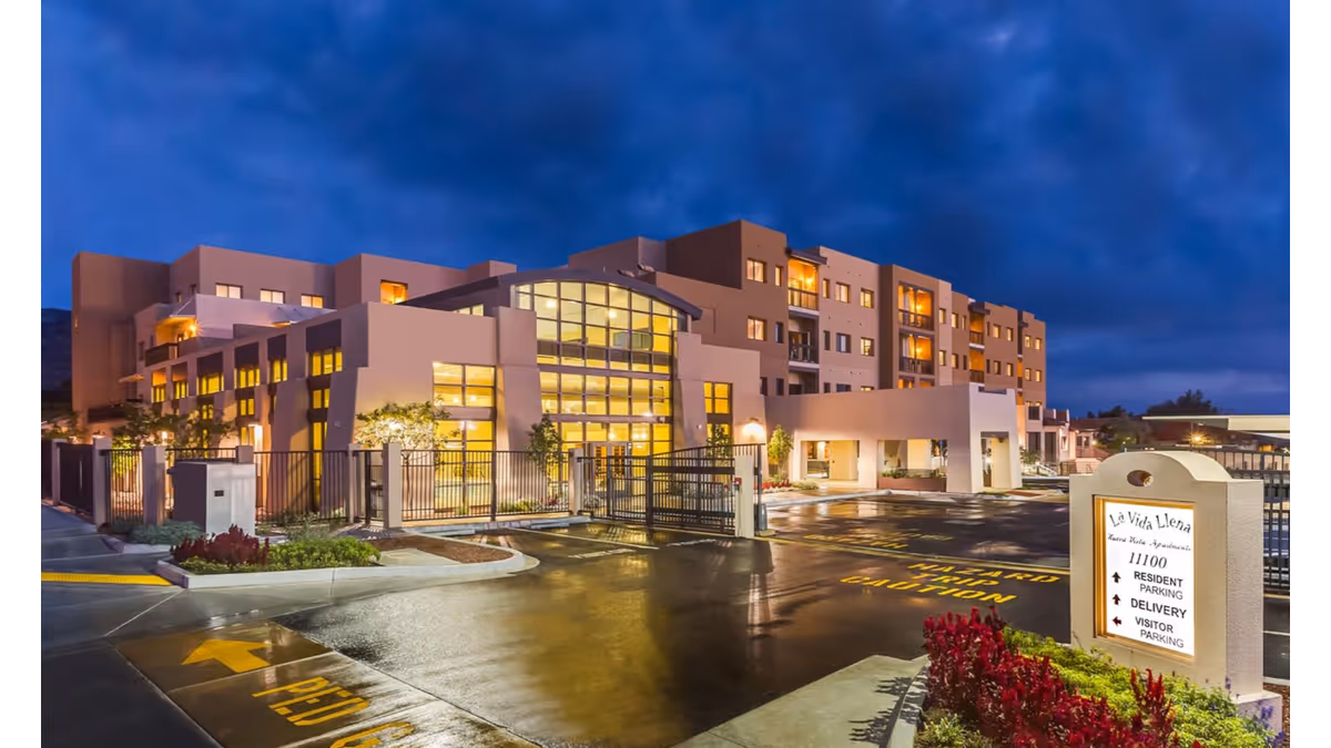 Exterior view of La Vida Llena Life Plan Retirement Community building at dusk with lights glowing from inside. The multi-story building features large windows and a gated entrance. A sign near the entrance indicates directions for resident parking, delivery, and visitor parking. The sky is cloudy and dark blue.