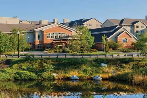 Front exterior of Brooksby Village senior living community with brick buildings, pond, and trees.