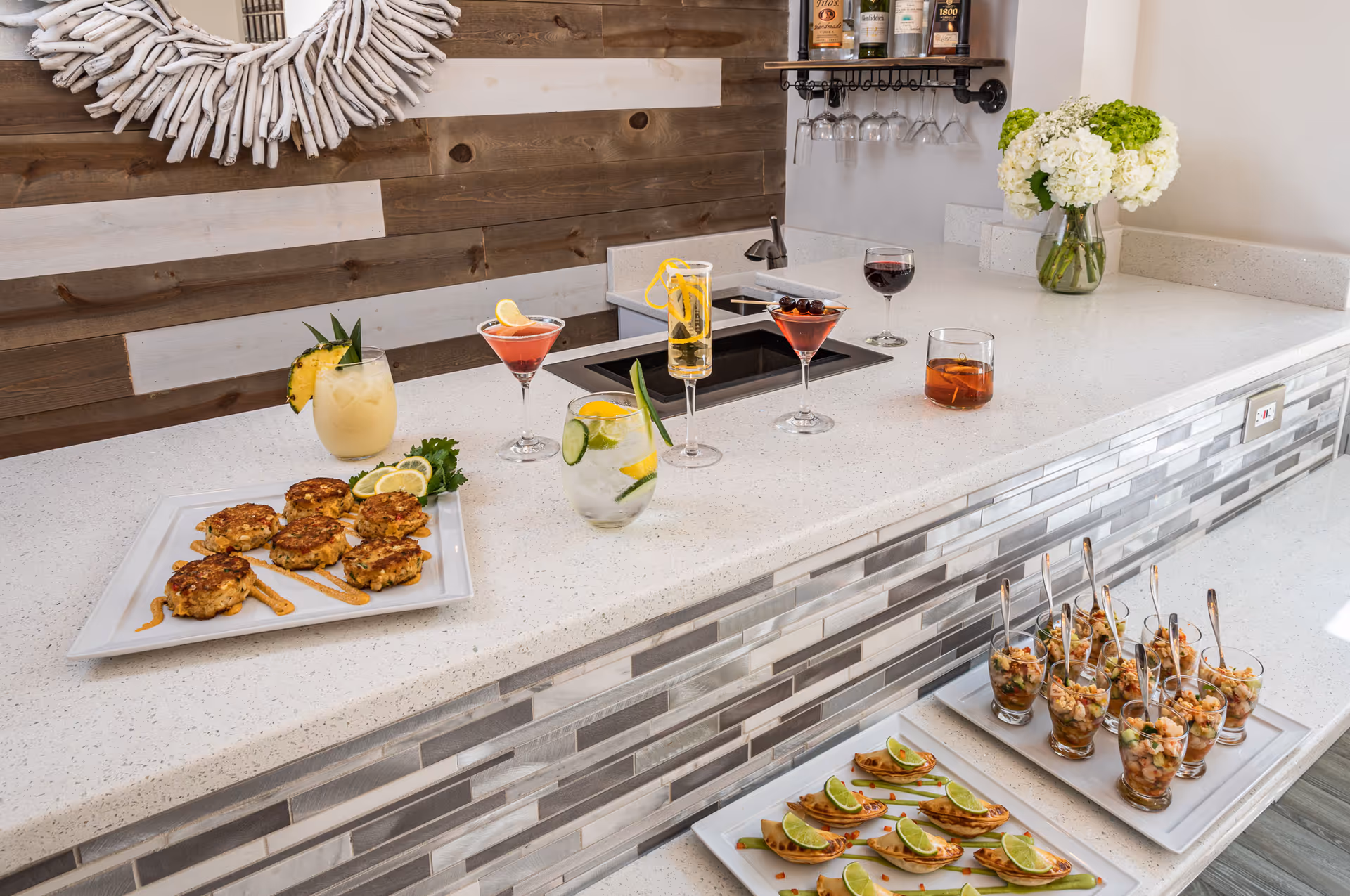 A modern kitchen bar area with a white countertop and a tiled front. On the counter are various cocktails and appetizers, including crab cakes, shrimp ceviche in small glasses, and lime-topped empanadas. There is a wooden wall with a decorative white wreath and a shelf holding bottles and hanging wine glasses. A vase with white and green flowers is also on the counter.