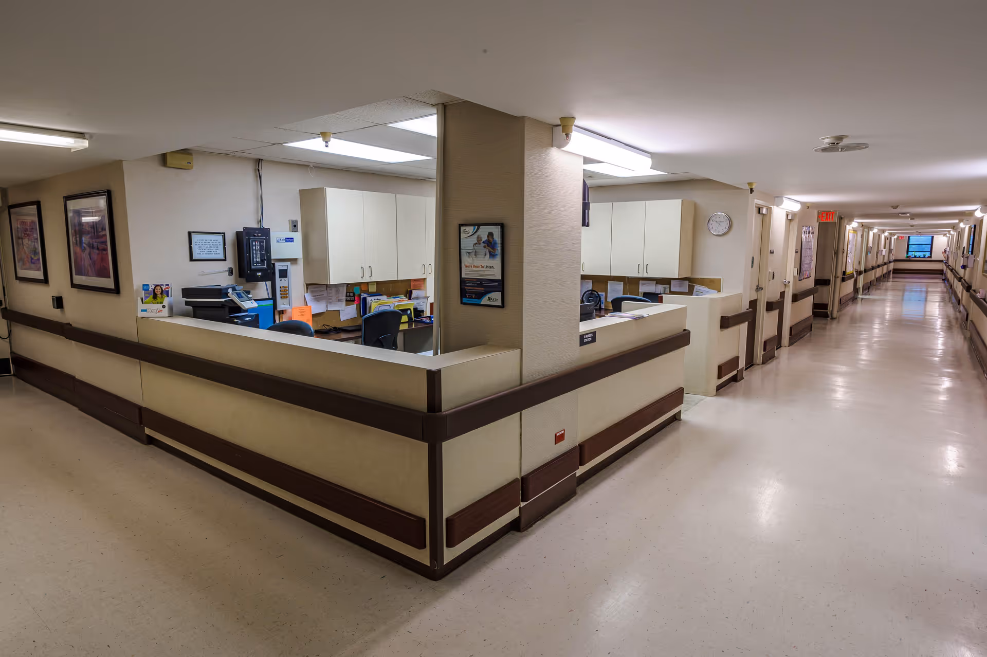 Interior view of a healthcare facility hallway with a nurse's station on the left side. The nurse's station has desks, chairs, cabinets, office equipment, and paperwork. The hallway is long with handrails on both sides and doors leading to rooms. The area is well-lit with ceiling lights and has a clean, polished floor.