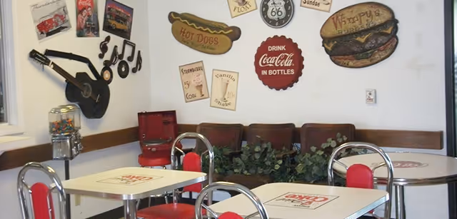 Interior of a retro-style dining area with tables and chairs featuring Coca-Cola branding. The walls are decorated with vintage signs including a guitar, musical notes, hot dogs, Route 66, and a hamburger. There is a gumball machine and a red jukebox in the corner.