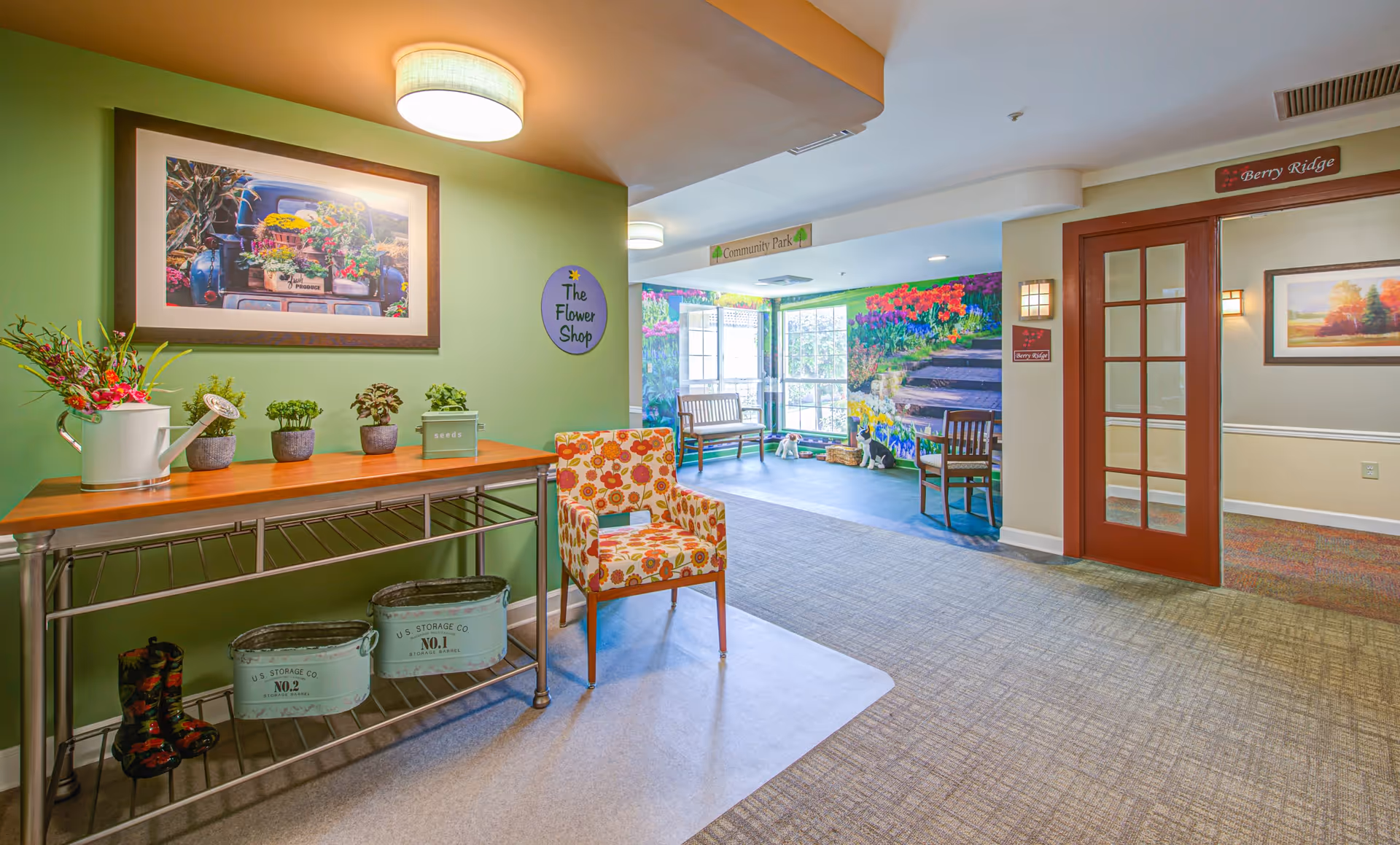 A bright and welcoming interior space in a senior living community featuring a green accent wall with a framed picture of a flower-filled truck and a sign that reads 'The Flower Shop'. Below the picture is a metal table with a wooden top holding small potted plants and a watering can. A colorful floral-patterned chair is placed nearby on a light rug. In the background, there is a seating area with wooden chairs and a bench near large windows, with a vibrant mural of flowers and a garden path on the wall. To the right, there are double glass doors with a sign above that reads 'Berry Ridge'.