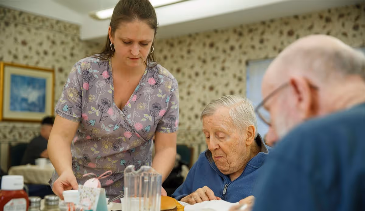 A caregiver in a floral patterned scrub top assists an elderly man seated at a table with food in a dining room. Another elderly man is partially visible in the foreground. The room has floral wallpaper and framed artwork on the wall.