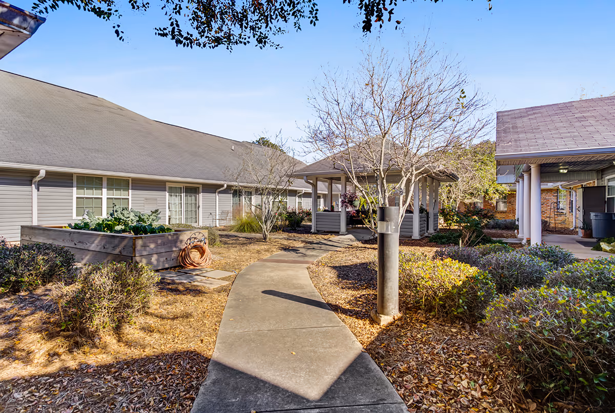 Outdoor courtyard area of a senior living facility with a concrete pathway leading to a covered gazebo surrounded by bushes, trees, and garden beds. The buildings have light gray siding and multiple windows, with a clear blue sky overhead.