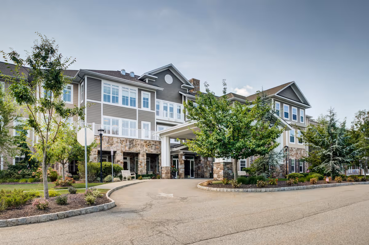 Front entrance and facade of a multi-story senior living building with a covered porte-cochère, trees, and landscaped driveway.