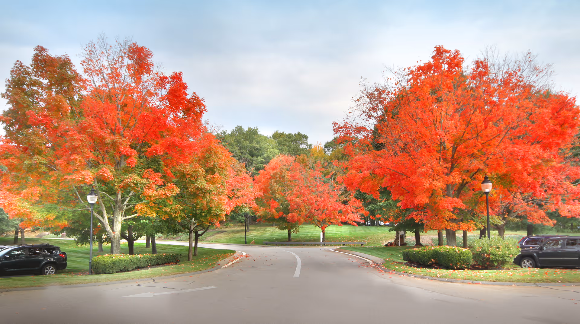 A paved road lined with vibrant red and orange autumn trees on both sides, with green grass and parked cars visible. The sky is partly cloudy.