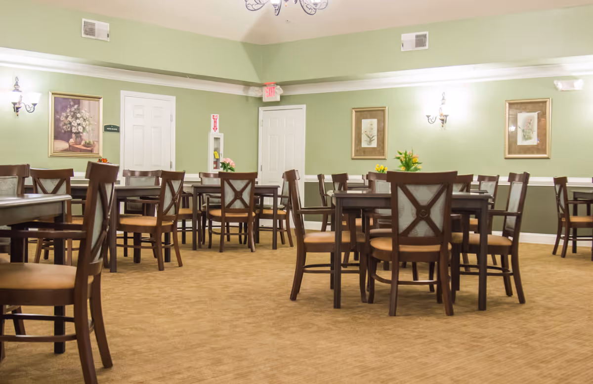 A dining room with multiple wooden tables and chairs arranged neatly on a beige carpet. The walls are painted light green with white trim, decorated with framed floral artwork and wall sconces. There are two white doors and an exit sign above one door.