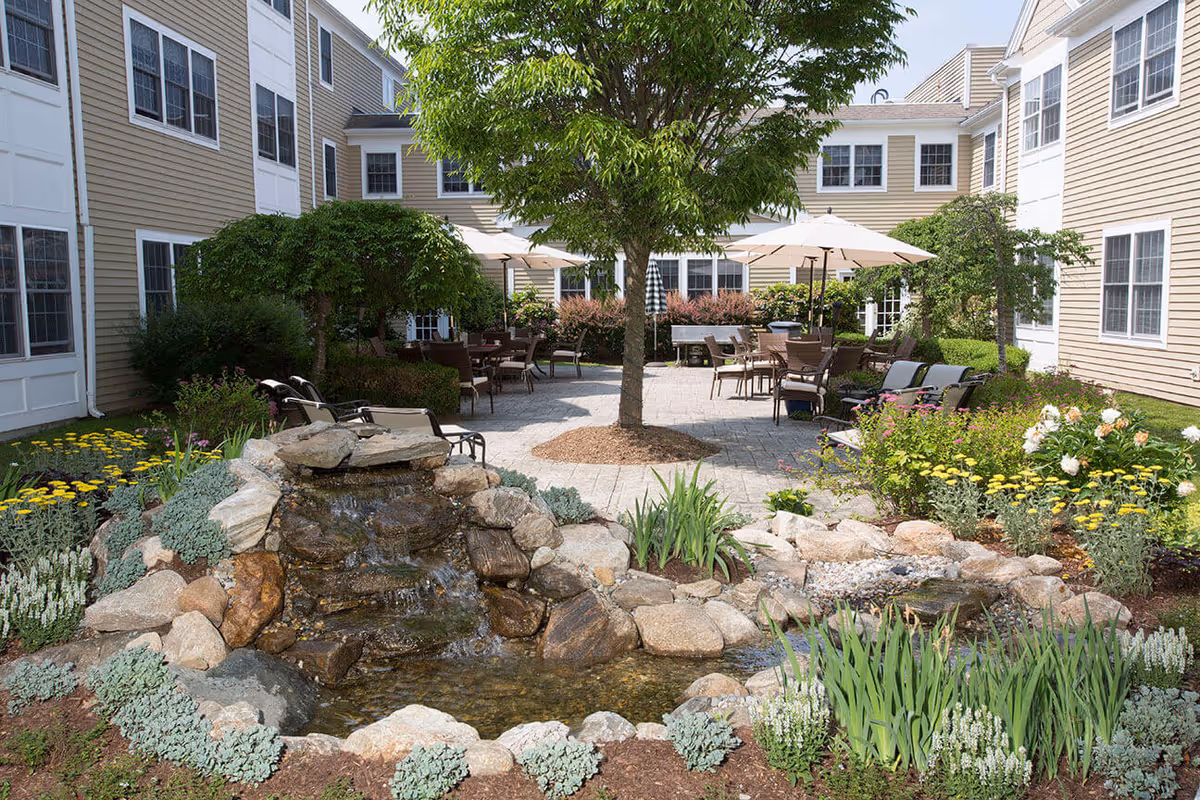 Outdoor courtyard area at Benchmark at Ridgefield Crossings featuring a small rock waterfall pond surrounded by various plants and flowers. The courtyard has multiple seating areas with tables, chairs, and umbrellas, shaded by a large tree in the center. The surrounding building has beige siding with white trim and multiple windows.