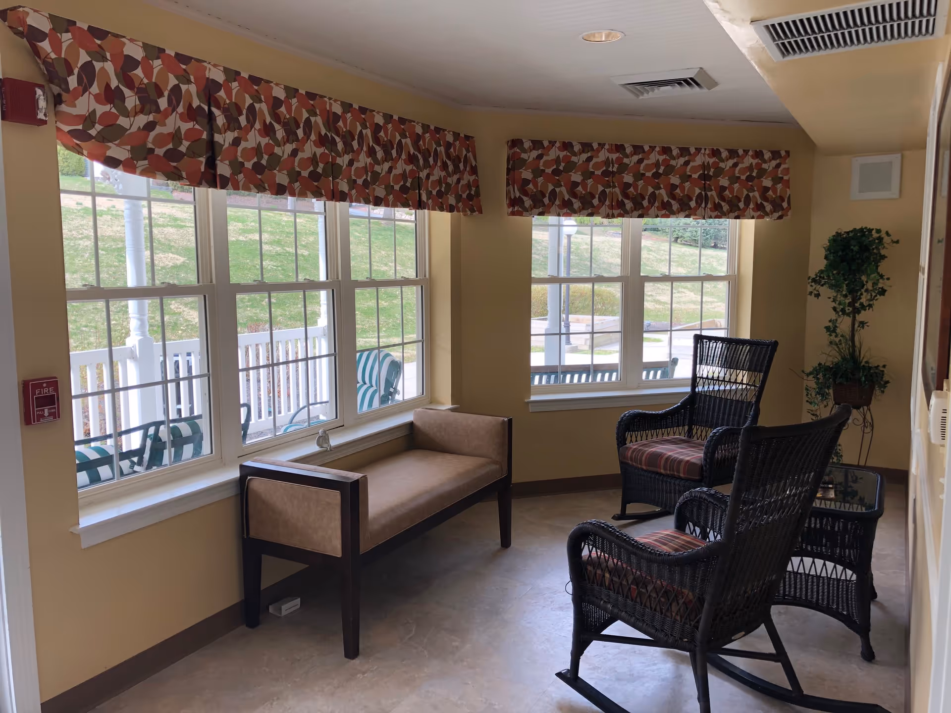 A cozy sitting area with two black wicker rocking chairs with striped cushions, a small glass-top wicker table, and a beige cushioned bench. The room has large windows with patterned valances, allowing natural light to brighten the space. Outside, green grass and patio furniture are visible.