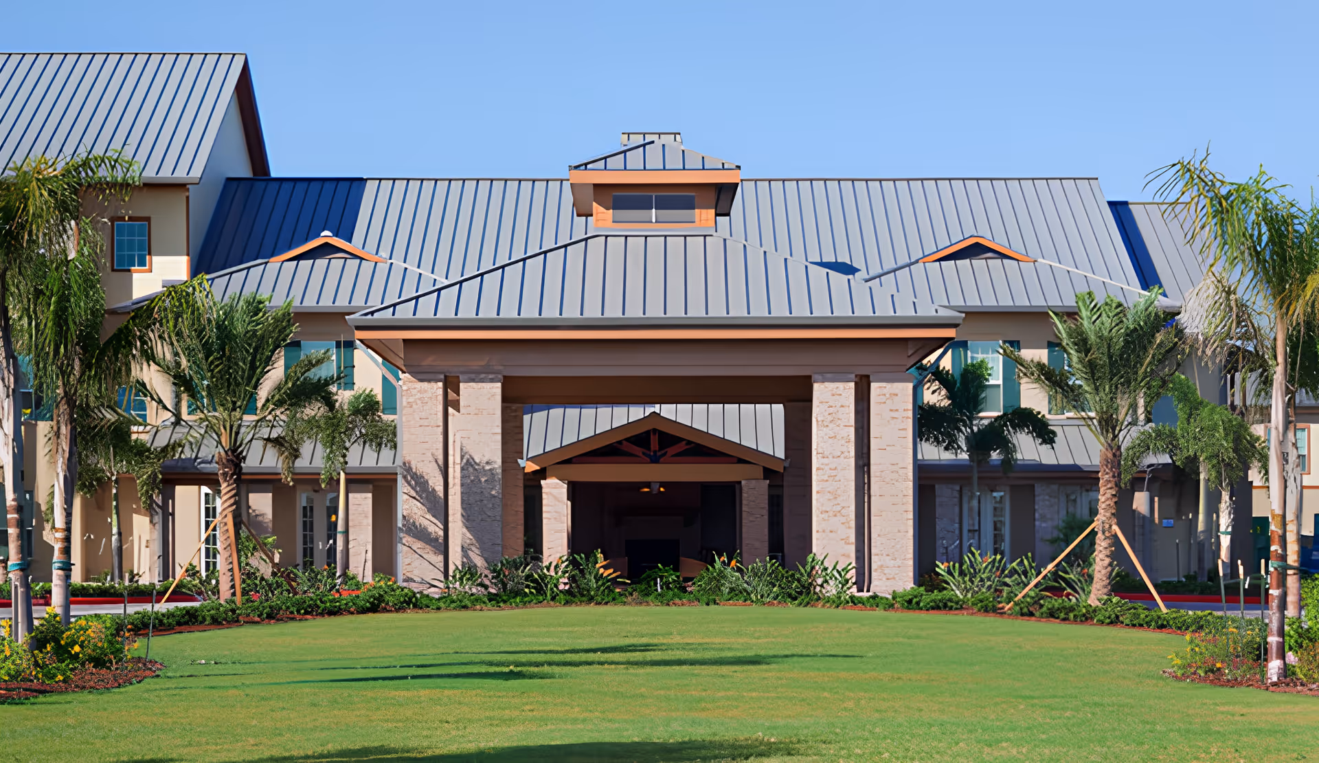 Front exterior view of Mirador Retirement Community building with a large covered entrance, surrounded by palm trees and well-maintained green lawn under a clear blue sky.