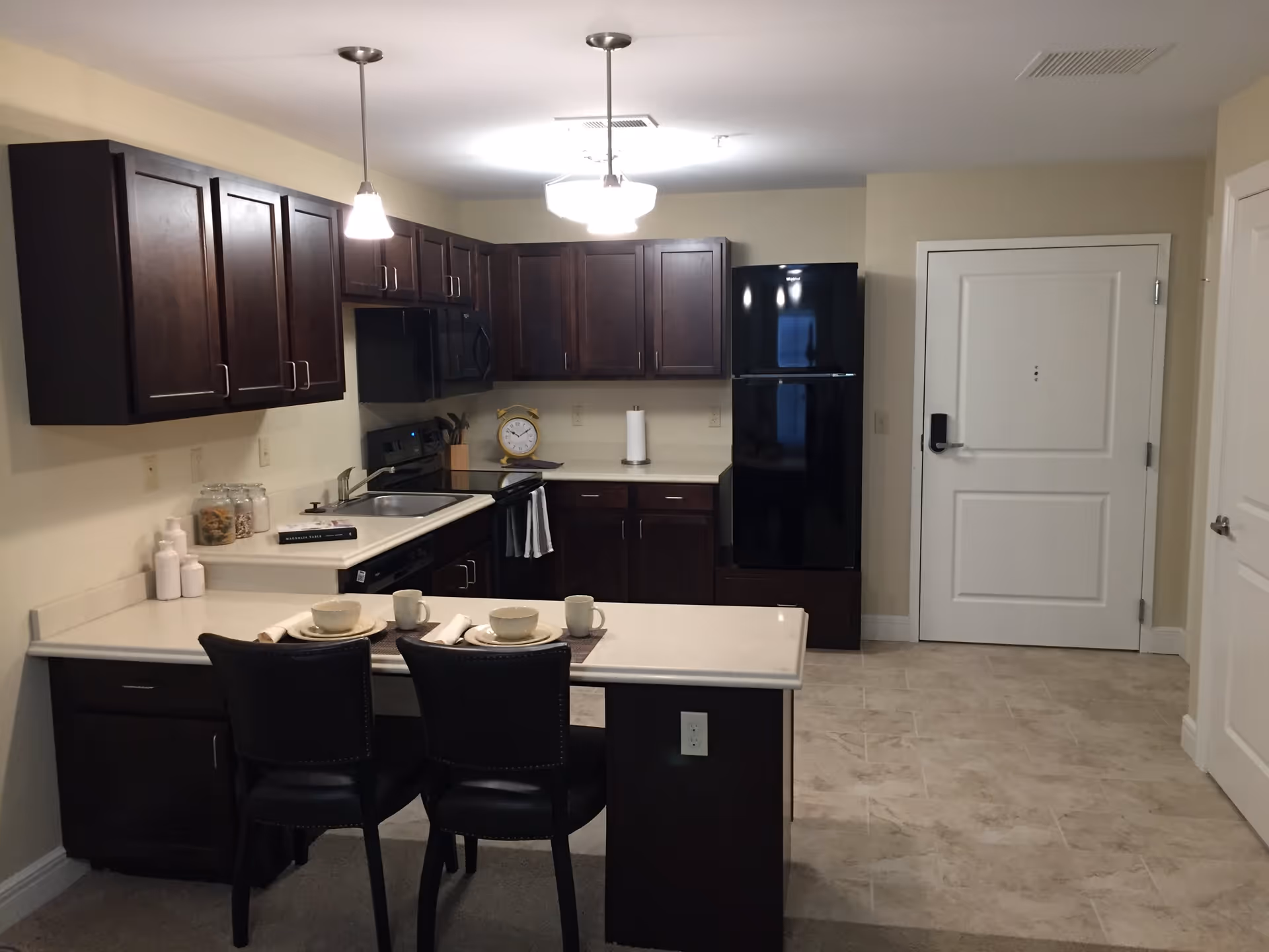Modern kitchen with dark wood cabinets, a black refrigerator, stove, microwave, and sink. There is a white countertop with two place settings and two black chairs. The floor is tiled near the refrigerator and door, with carpet near the seating area. Two pendant lights hang from the ceiling.