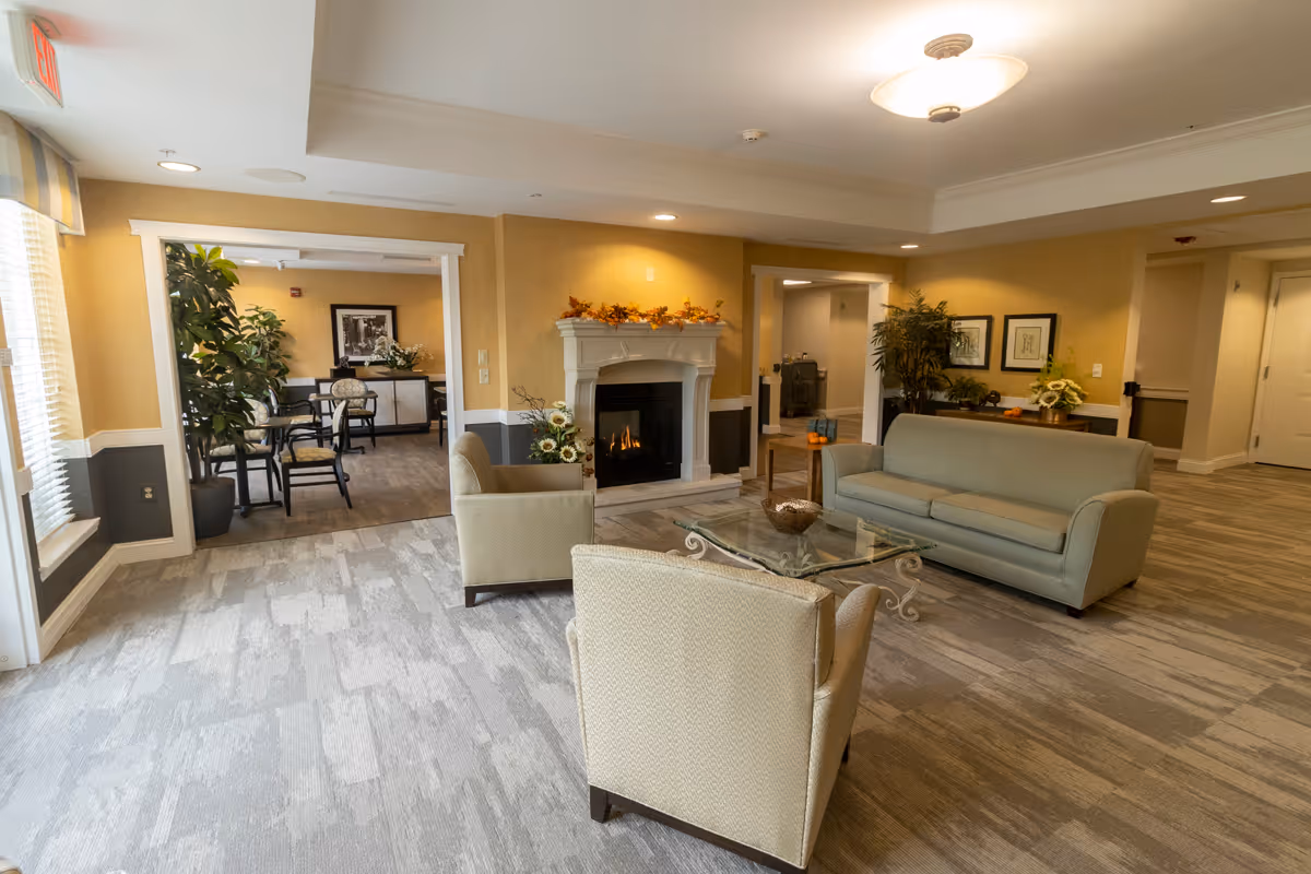 A cozy living room area in a senior living facility with a light green sofa, two beige armchairs, and a glass coffee table. There is a fireplace with a mantle decorated with autumn leaves and flowers. The room has soft yellow walls with white trim, recessed lighting, and a large window with blinds. In the background, there is a dining area with tables and chairs, plants, and framed artwork on the walls.