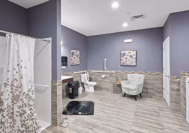 A spacious bathroom with purple walls and tiled floor. The room features a white toilet with grab bars, a shower with a white curtain decorated with a leaf pattern, a black trash bin, a small table with a potted plant, and a floral upholstered armchair. Two framed floral artworks hang on the walls.