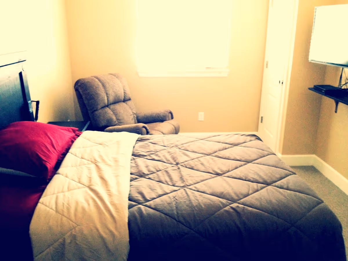 A bedroom with a neatly made bed featuring a gray quilt and red pillows. There is a cushioned armchair next to a window with blinds, a small nightstand beside the bed, and a wall-mounted TV on the right side.