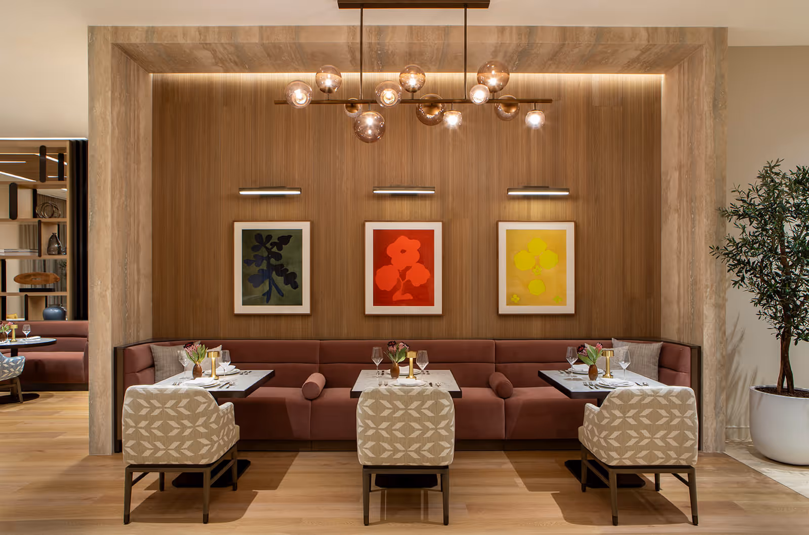 A modern dining area with three tables set for two, featuring beige patterned chairs and a long maroon cushioned bench against a wooden paneled wall. Above the bench are three framed abstract floral artworks in green, red, and yellow, each illuminated by individual wall lights. A contemporary chandelier with multiple glass globes hangs from the ceiling, and a large potted plant is visible on the right side.