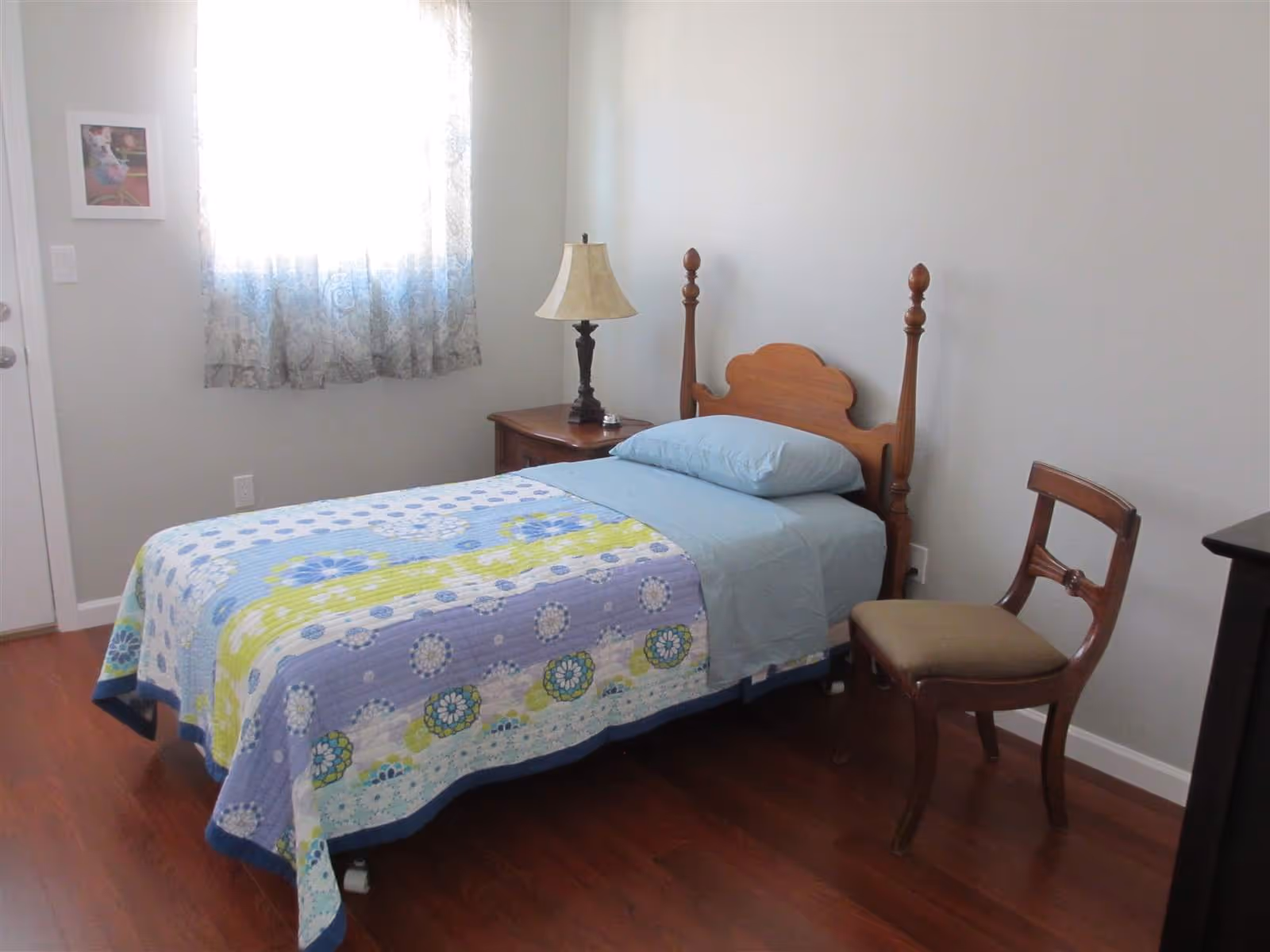 A small bedroom with a single wooden bed covered with a colorful quilt and blue sheets. Next to the bed is a wooden nightstand with a lamp and a small bell. A wooden chair with a cushioned seat is placed beside the bed. A window with sheer curtains allows natural light into the room, and a framed picture hangs on the wall near the window.