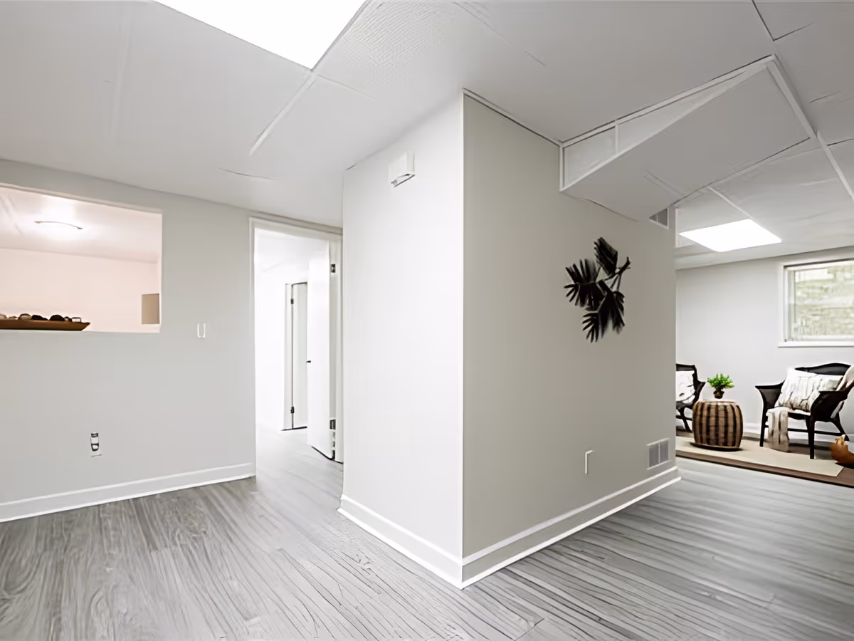 Interior view of a modern hallway with light gray walls and wood-patterned flooring. The hallway leads to a small sitting area with two chairs, a small table with a plant, and a window letting in natural light. A decorative wall piece with black leaves is mounted on the corner wall.