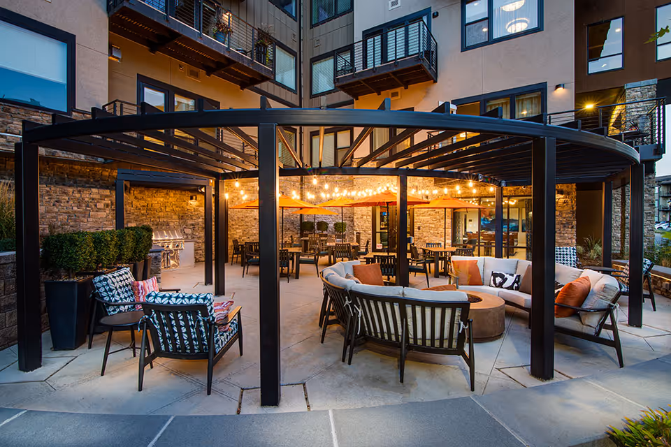 Covered outdoor courtyard with sofas, chairs, dining tables, string lights and umbrellas in front of a multi-story building.