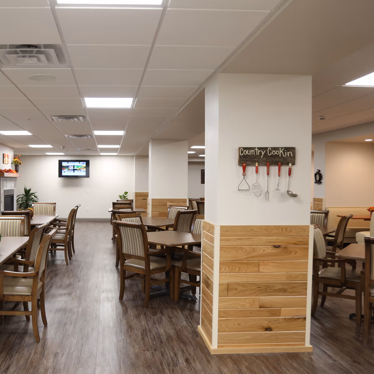 A spacious dining room with multiple wooden tables and chairs arranged neatly on a wood floor. The walls are light-colored with wood paneling on the lower half. A pillar in the center has a decorative sign that reads 'Country Cookin' with kitchen utensils hanging below it. A flat-screen TV is mounted on the far wall above a fireplace with plants and decorations nearby.