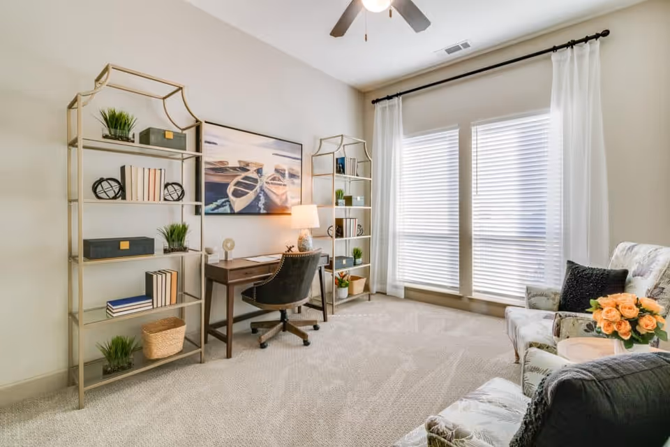 Bright living room with a desk and chair flanked by bookshelves, large windows with sheer curtains, and upholstered seating with a side table and flowers.